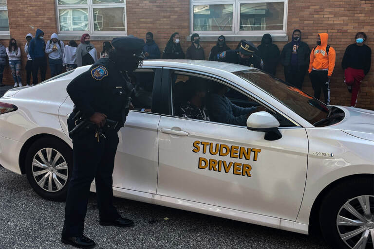 A Wilmington police officer approaches a car with teen drivers during a simulation witnessed by other students at William Penn High School. (Cris Barrish/WHYY)