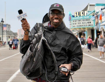 A reporter poses on a beach boardwalk with his recording equipment, smiling and holding his microphone aloft.