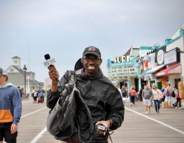 A reporter poses on a beach boardwalk with his recording equipment, smiling and holding his microphone aloft.