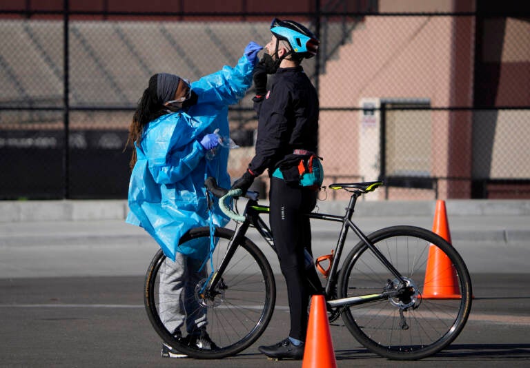 A medical technician performs a nasal swab test on a cyclist queued up in a line with motorists at a COVID-19 testing site near All City Stadium Thursday, Dec. 30, 2021, in southeast Denver. With the rapid spread of the omicron variant paired with the Christmas holiday, testing sites have been strained to meet demand both in Colorado as well as across the country