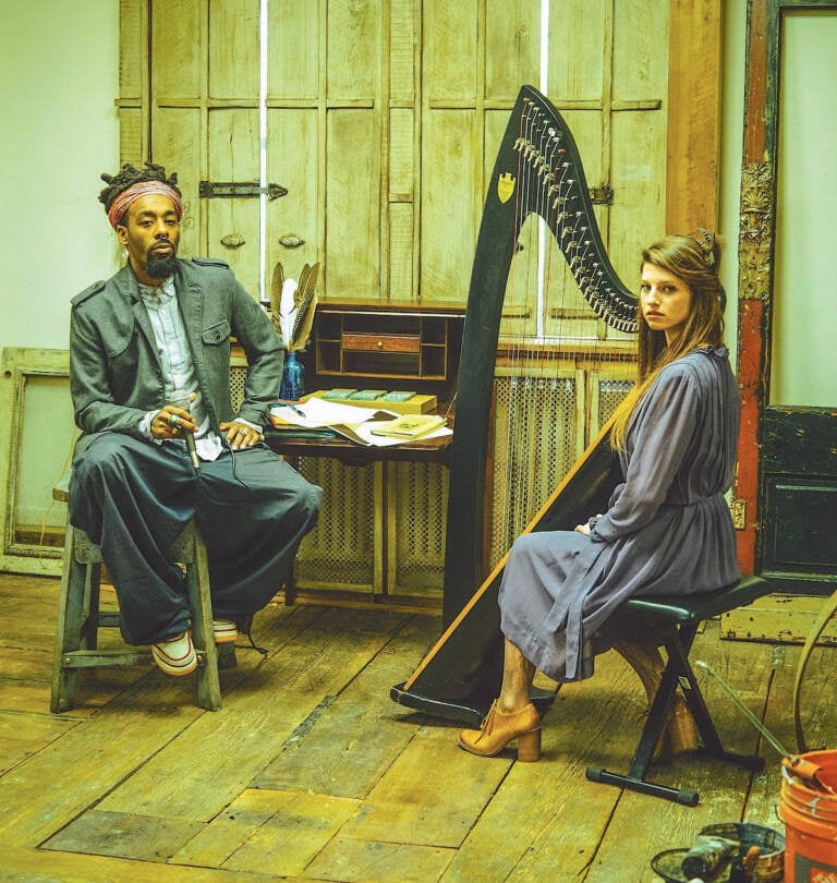 Kuf Knotz sits while holding a mic, as Christine sits next to her harp