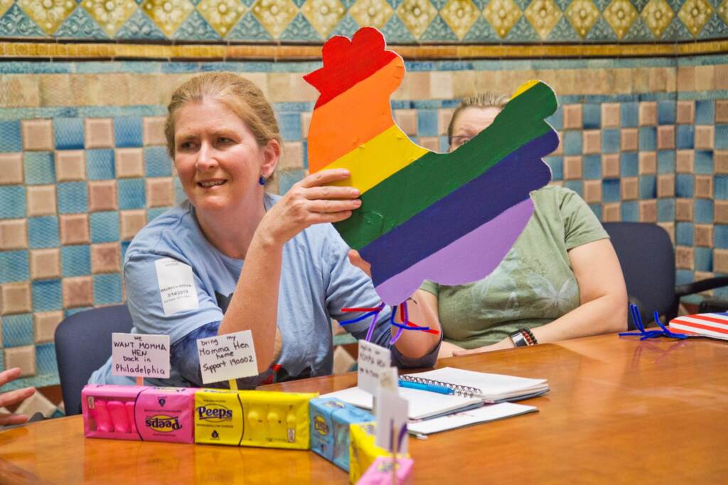 Maureen Breen sits in front of a table with signs advocating for legal hens in Philadelphia. She holds a cardboard sign in the shape of a hen in rainbow colors. 