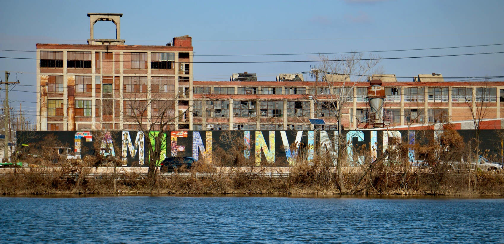 With the backdrop of a abandoned factory, the ''Camden Invincible'' mural faces Admiral Wilson Boulevard and the Cooper River. (Emma Lee/WHYY)