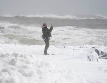 Waves crash behind a man as he uses a cell phone to capture images on a snow-covered beach, Saturday, Jan. 29, 2022.(AP Photo/Julio Cortez)