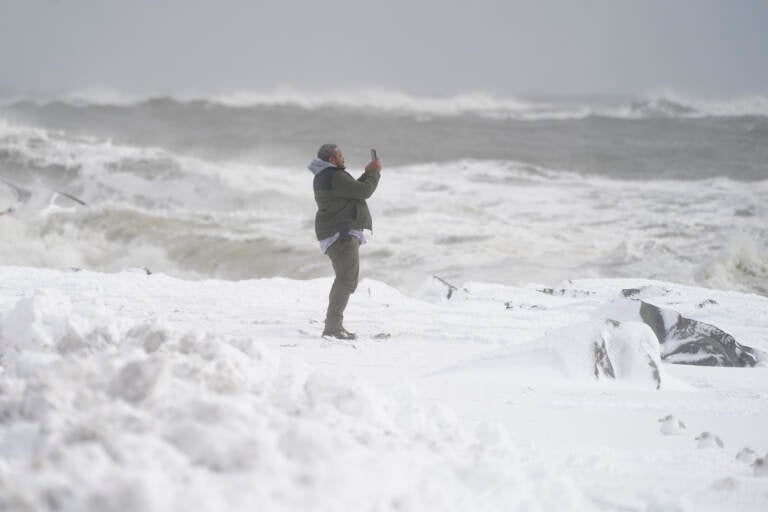 Waves crash behind a man as he uses a cell phone to capture images on a snow-covered beach, Saturday, Jan. 29, 2022.(AP Photo/Julio Cortez)