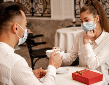 Young couple in medical masks clinking cups of tea near gift box on table in cafe