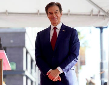 Mehmet Oz stands with hands clasped in front of him, wearing a dark blue suit and a red tie.