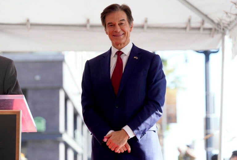 Mehmet Oz stands with hands clasped in front of him, wearing a dark blue suit and a red tie.