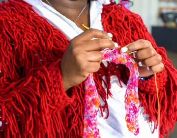 A close up of Emani Outterbridge crocheting a pink, orange, and white piece
