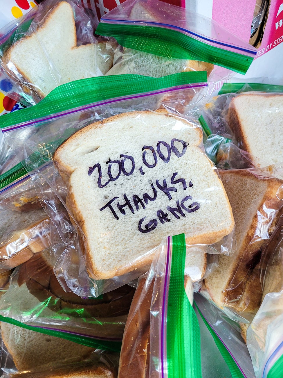 A pile of peanut butter and jelly sandwiches packed in zip lock bags. The top sandwich has "200,000 THANKS GUYS" written on the back.