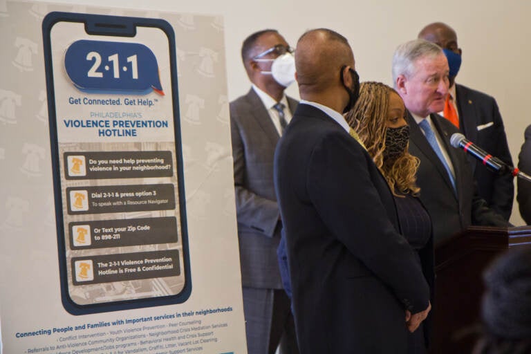 A sign announcing the gun violence prevention hotline is shown in the foreground, with elected officials speaking at a podium in the background.