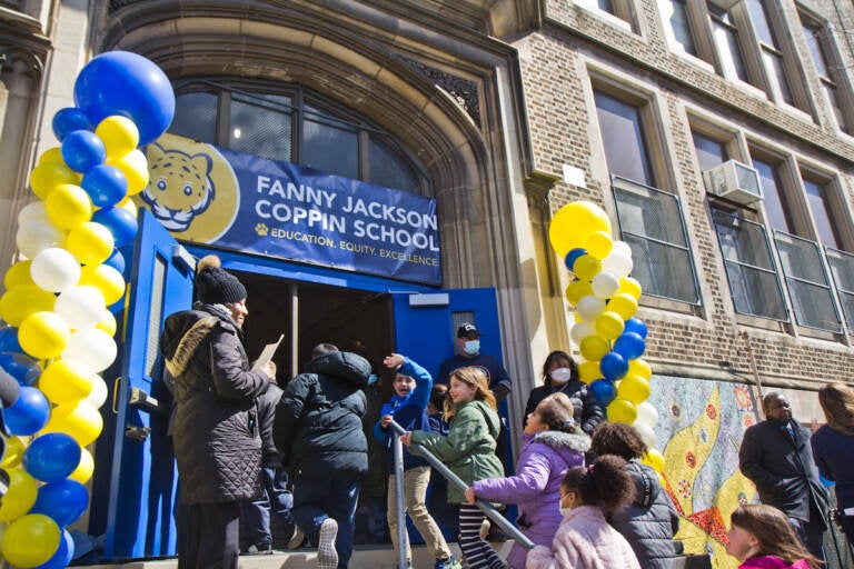Children are running toward the open doorway of a school surrounded by white, yellow, and blue balloons.