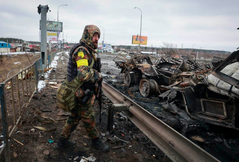 An armed man stands by the remains of a Russian military vehicle in Bucha, close to the capital Kyiv, Ukraine, Tuesday, March 1, 2022. Russia on Tuesday stepped up shelling of Kharkiv, Ukraine's second-largest city, pounding civilian targets there. Casualties mounted and reports emerged that more than 70 Ukrainian soldiers were killed after Russian artillery recently hit a military base in Okhtyrka, a city between Kharkiv and Kyiv, the capital.