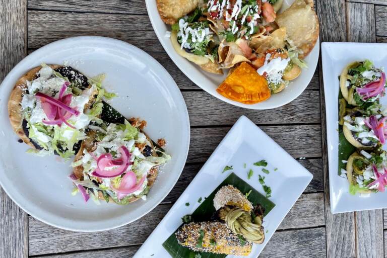 A spread of empanadas, elote, tostadas and more on white plates are shown from above at Las Cazuelas Restaurant, with a wooden table in the background.