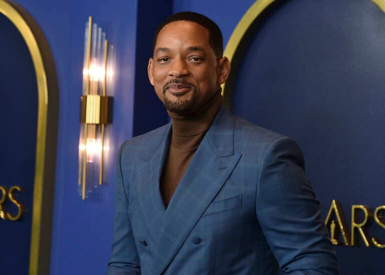 Will Smith, wearing a brown turtleneck and a dark blue plaid suit jacket, is posing in front of a dark blue background at the 94th Academy Awards nominees luncheon on Monday, March 7, 2022, in Los Angeles.