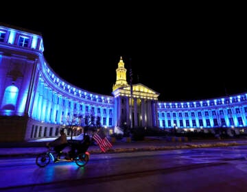 An American flag unfurls off a pedicab as it glides past the Denver City/County Building, which is illuminated in yellow and blue in support of Ukraine, late Monday, Feb. 28, 2022, in Denver. (AP Photo/David Zalubowski)