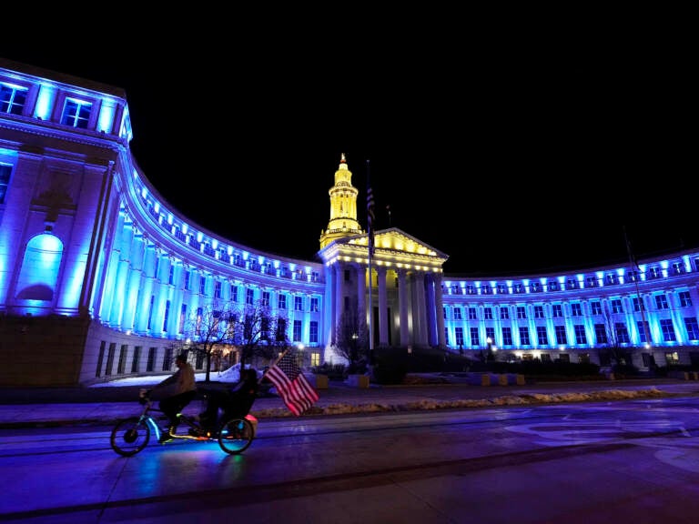 An American flag unfurls off a pedicab as it glides past the Denver City/County Building, which is illuminated in yellow and blue in support of Ukraine, late Monday, Feb. 28, 2022, in Denver. (AP Photo/David Zalubowski)