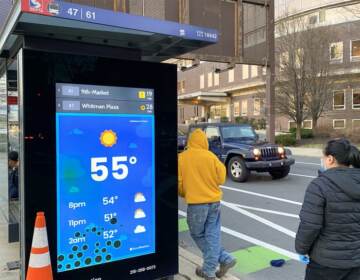 People stand at a SEPTA bus stop which has a screen, displaying the weather, temperature, and real-time arrival information for buses.