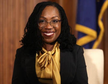 A portrait-style photo of U.S. Supreme Court nominee, Ketanji Brown Jackson. She's smiling, wearing a gold-colored blouse and black jacket.