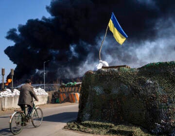 A man rides a bicycle past a structure with the Ukrainian flag flying on top, as smoke billows in the background.