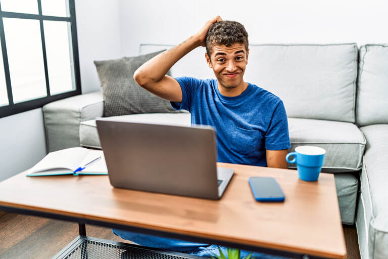 A young man sitting on the floor using a laptop with a hand on his head looking confused and uncertain.