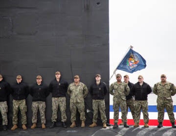 The crew of the USS Delaware stand in front of a flag of the Presidental Seal while on their submarine during the dress rehearsal for the official commission ceremony for the new submarine.