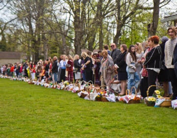 A row of Ukrainian American parishioners stand on the edge of a green field, with Easter baskets in front of them.