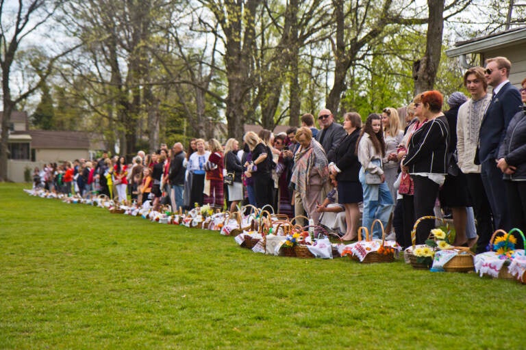 A row of Ukrainian American parishioners stand on the edge of a green field, with Easter baskets in front of them.