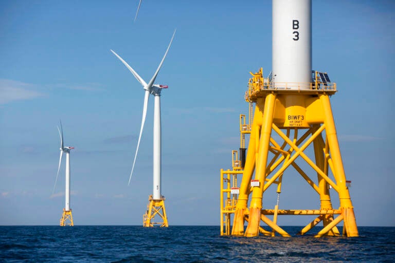 Wind turbines are seen up close and in the distance in the ocean.
