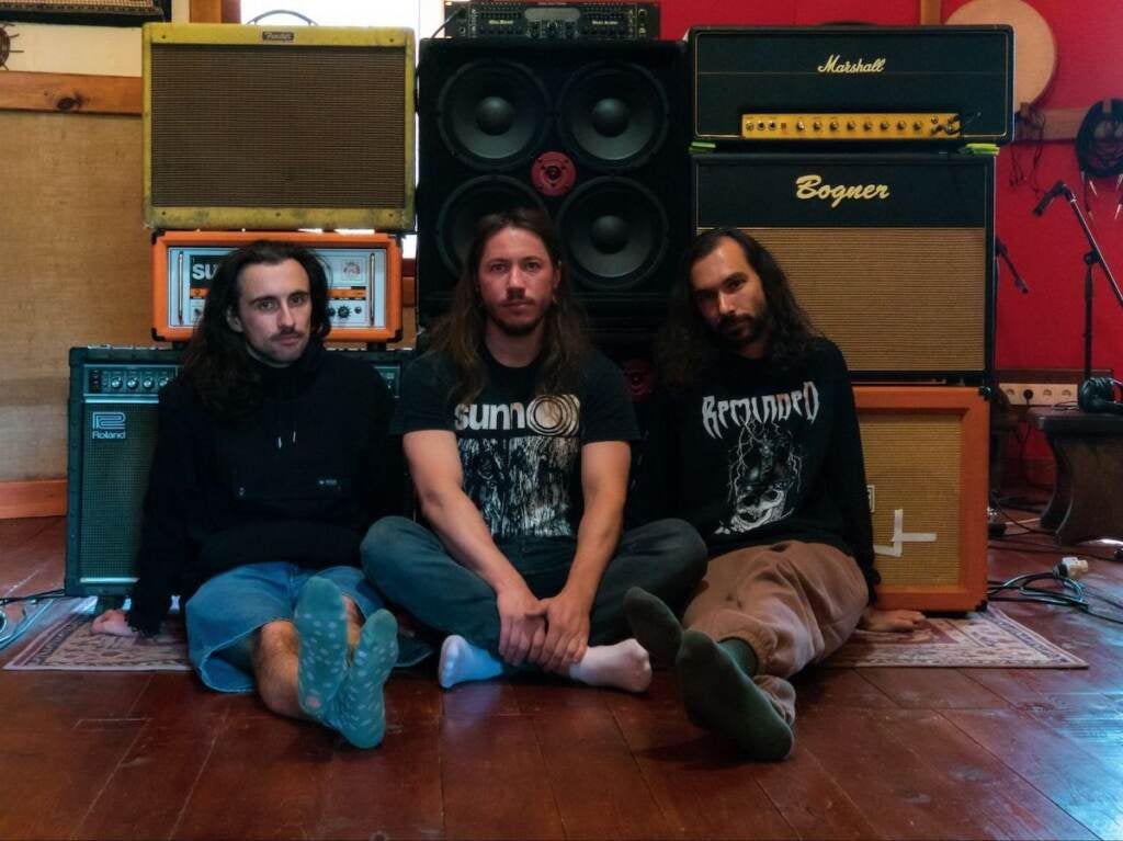 The band members of the Ukrainian punk band Кат sit on a hardwood floor, with music amps behind them.