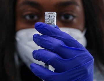 A medic wearing a facemask and dark blue gloves holds up a vaccine container.