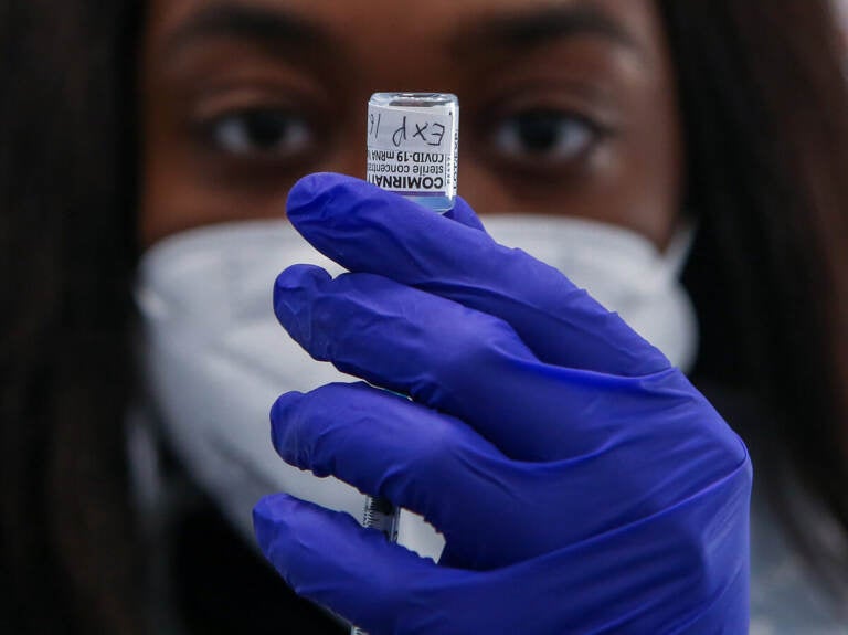 A medic wearing a facemask and dark blue gloves holds up a vaccine container.