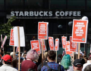 A Starbucks coffee shop is seen in the background as people protest against the company, holding signs in support of unionizing staffers