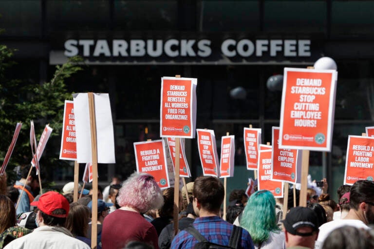 A Starbucks coffee shop is seen in the background as people protest against the company, holding signs in support of unionizing staffers