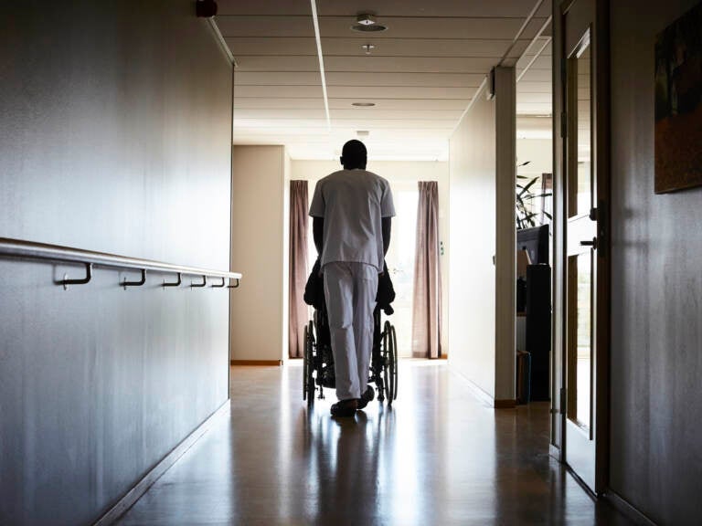 A person dressed in scrubs pushes someone in a wheelchair down a hallway.