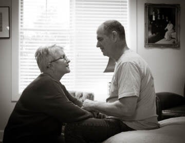 Mary Daniel kneels down by her husband, seated on a bed at his Florida memory care facility.