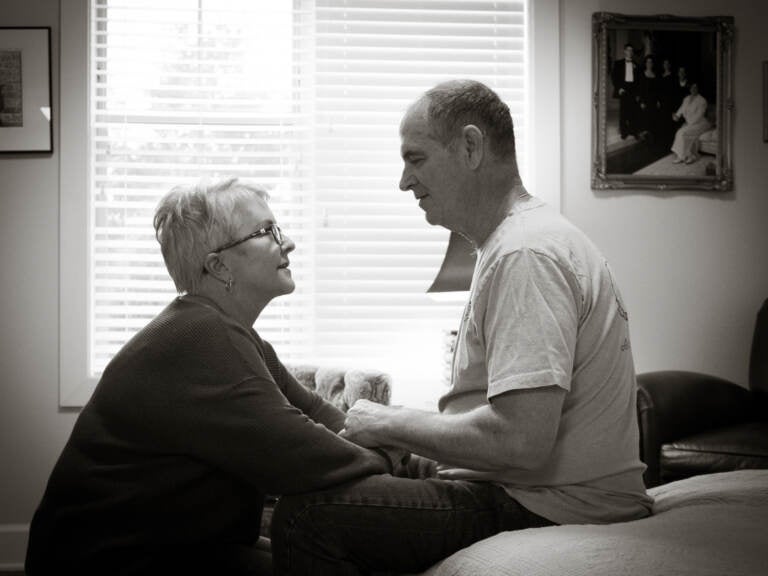 Mary Daniel kneels down by her husband, seated on a bed at his Florida memory care facility.