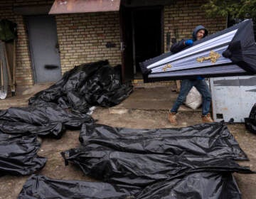 A man carries a coffin next to plastic bags with corpses of civilians