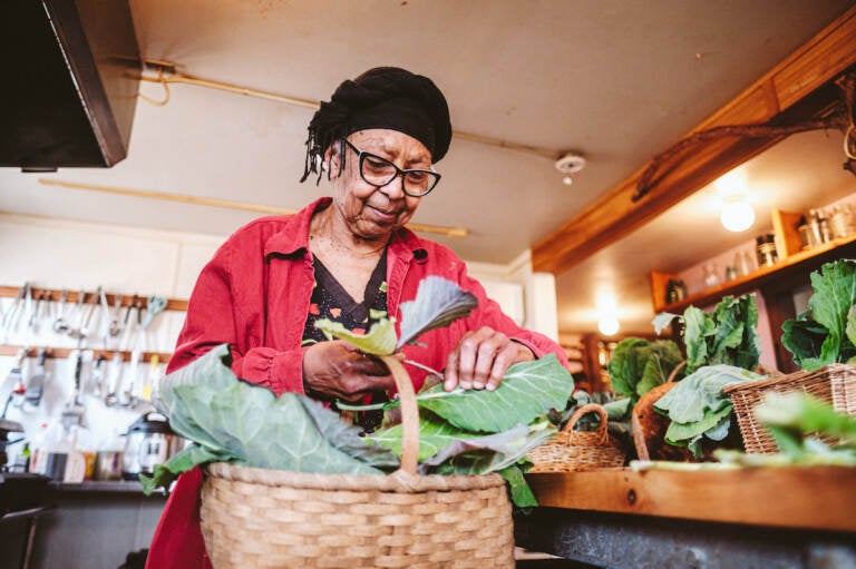 Ira Wallace, wearing a red jacket, takes a piece of collard greens in the basket before her.