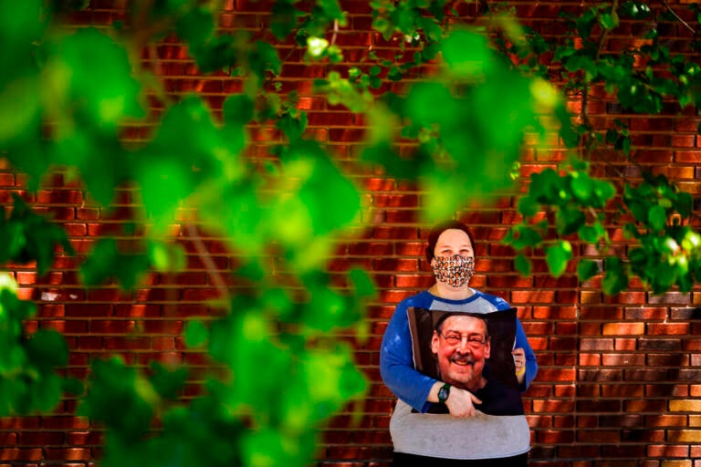 Sara Atkins poses for a photo, in Wynnewood, Pa., Tuesday, May 10, 2022, while holding a pillow with an image of her father Andy Rotman-Zaid, who died of COVID-19 in December 2020. She channels her grief into fighting for global vaccination and better access to health care to honor her father.