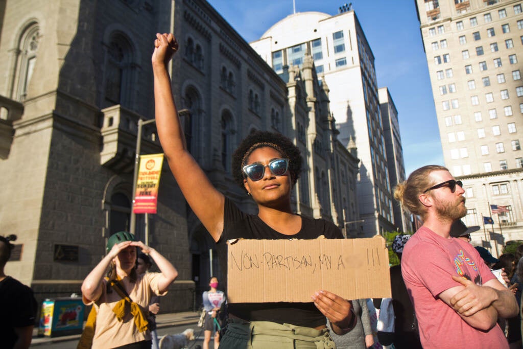 Kendall Bedford holds up a fist in the air while holding a protest sign that says ''Nonpartisan my a**''