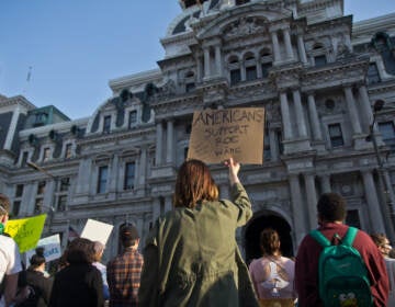 A person stands in a crowd of people in front of City Hall, holding a sign that says, 