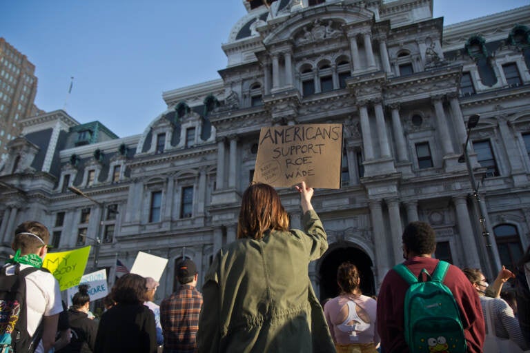 A person stands in a crowd of people in front of City Hall, holding a sign that says, 
