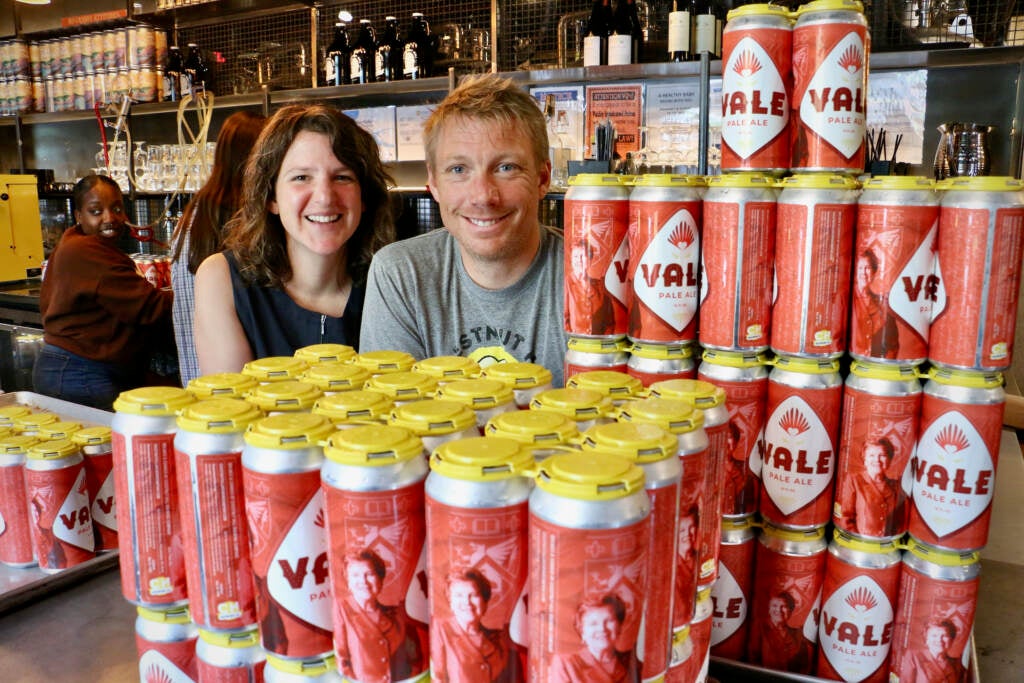 Co-owners of Chestnut Hill Brewing Company, Lindsey Pete and Nick Gunderson, look from behind a stack of beers that celebrate Chestnut Hill College's retiring president, Sister Carol Jean Vale.