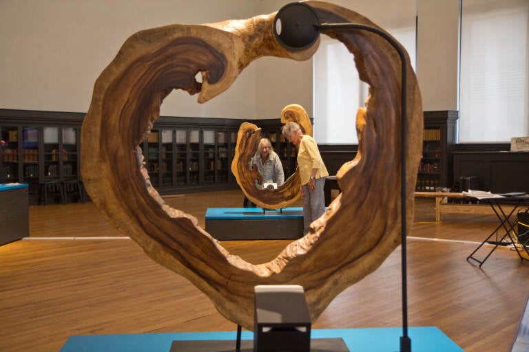 Two artists are visible in the background, with a wooden object from the exhibition in the foreground.
