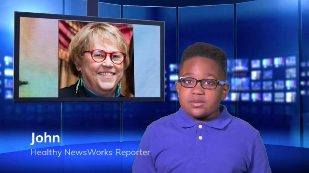 John, a Healthy NewsWorks reporter, faces the camera, with a screen with a person's face on it in the background.