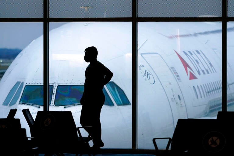 A silhouette of a person standing in an airport is visible, with a plane visible in the background, outside the window.