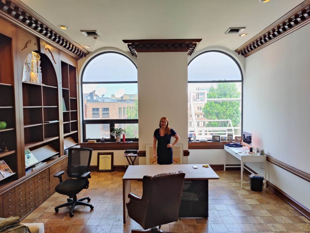 Alaine Arnott stands behind her desk in her office, with two light-filled windows behind her looking out onto Old City in Philadelphia.