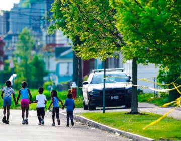 Children walk hand-in-hand down a tree-lined street, with yellow crime tape visible in the corner.