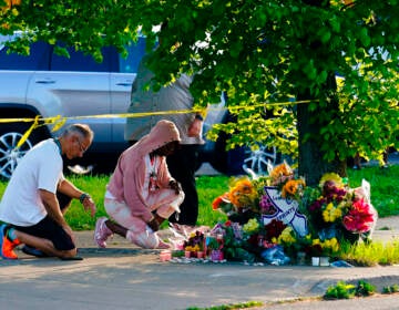 Two people kneel on the ground in front of a memorial made of flowers.